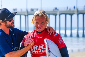 Westcliff Warriors Surf Team celebrating victory at Huntington Beach Pier after NSSA college surf competition, holding boards with Pacific waves in the background.